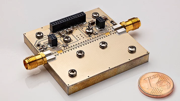 Close-up of a rectangular electronic circuit board featuring metallic connectors. Two gold-colored connectors are prominently positioned on opposite sides of the board. A one euro cent coin is placed for scale.
