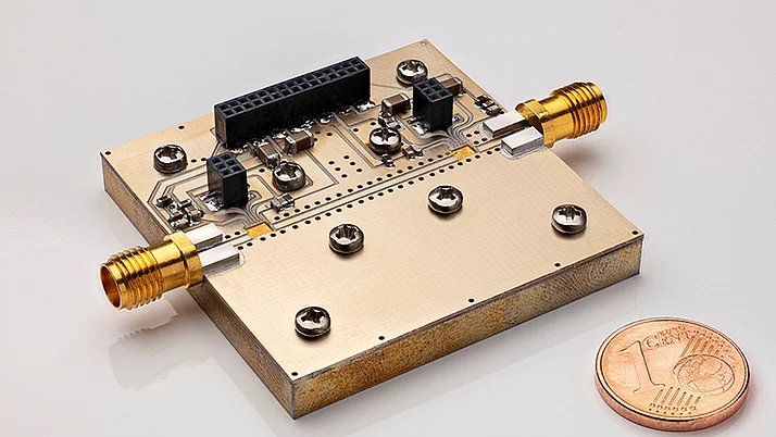 Close-up of a rectangular electronic circuit board featuring metallic connectors. Two gold-colored connectors are prominently positioned on opposite sides of the board. A one euro cent coin is placed for scale.