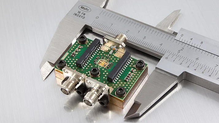Close-up of a green circuit board mounted on a metallic base, featuring multiple electronic components and connectors. A caliper is measuring the dimensions of the board.