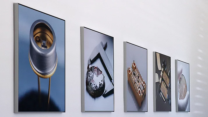 Fünf Bilder hängen an einer weißen Wand. Die Motive sind technische Objekte, darunter eine Drahtkammer, ein Messgerät und elektronische Schaltungen. Die Fotografien zeigen Details und Strukturen der Objekte in klaren, prägnanten Ansichten.