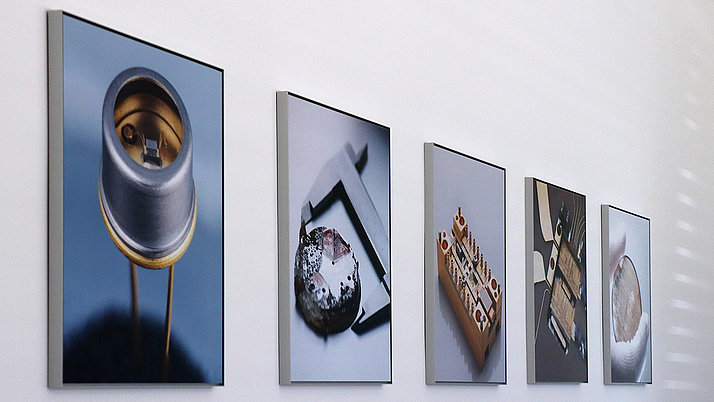 Five pictures hang on a white wall. The motifs are technical objects, including a wire chamber, a measuring device and electronic circuits. The photographs show details and structures of the objects in clear, concise views.
