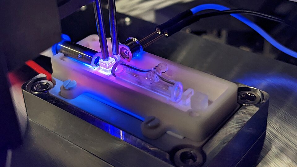 The image shows the integration of a miniaturized optic component on a ceramic micro-optical bench. The optic glows blue/violet due to the UV radiation used to cure the adhesive.