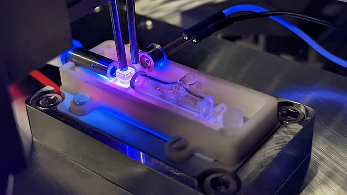 Die Abbildung zeigt die Integration einer miniaturisierten Optik auf einer mikrooptischen Bank aus Keramik. Duch die UV-Strahlung zum Aushärten des genutzten Klebstoffs leuchtet die Optik Blau/Violett.