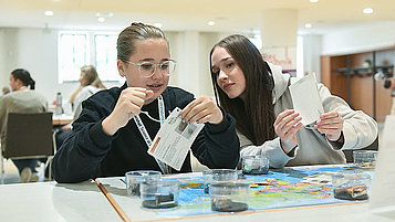 Two girls at a table with pieces of paper in their hands. On the table are a world map and various stones in glass bowls.