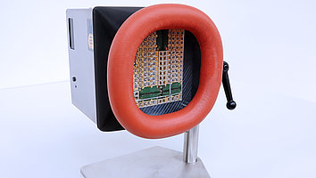 UV-LED-Bestrahlungssystem mit einer roten ovalen Aussparung, durch die eine kreisförmige Platine zu sehen ist. Das Gerät steht auf einem stabilen Metallfuß und hat einen Handhebel an der Seite. 