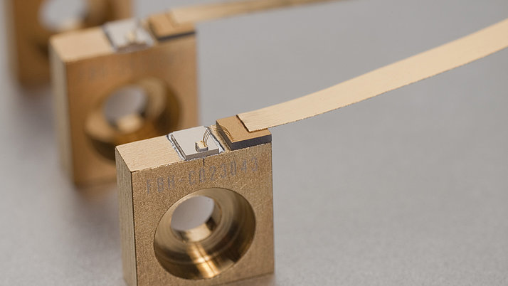 Three square, gold-colored metal pieces lie in a straight line on a neutral surface. Each piece has a hole in the middle and a thin, flat metal color that sticks out.