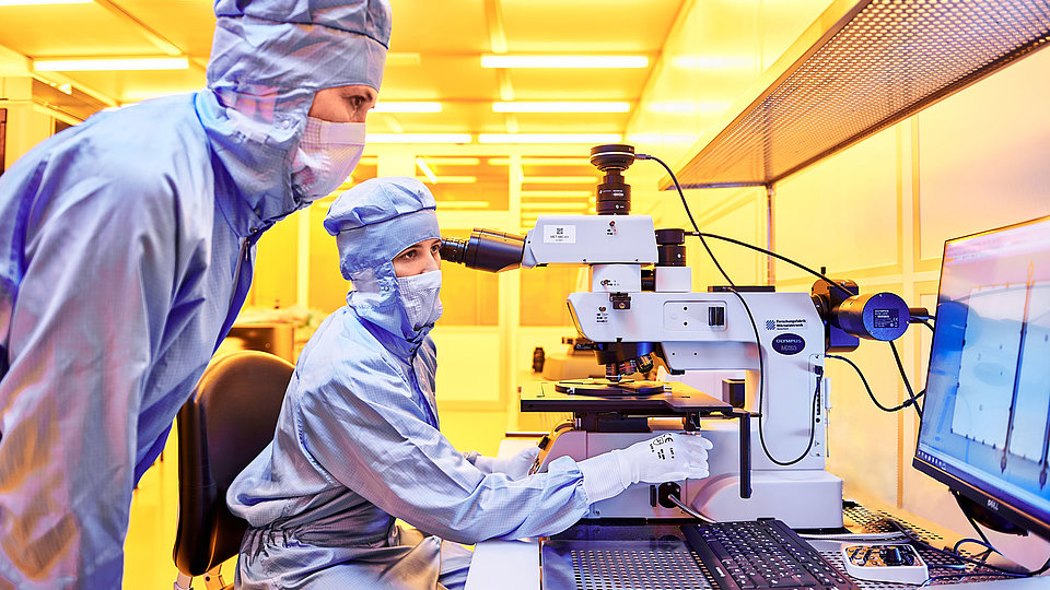 Two laboratory technicians in blue protective clothing observe a microscope. One technician is sitting and looking through the eyepiece, while the other stands beside, focused on the process.