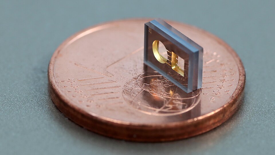 Tiny rectangular microchip standing upright on a one-cent coin, showing a gold circular element and internal structures, emphasizing its extremely small size relative to the coin.