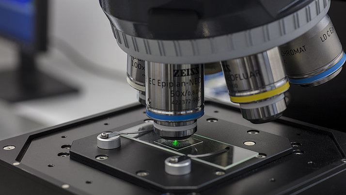 A microscope with several objectives is positioned on a sample. A green light illuminates the sample on the slide. 