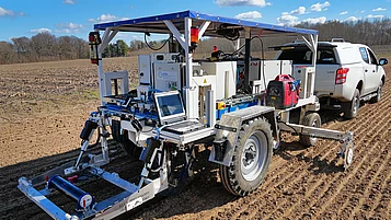 An agricultural vehicle with a sensor system is standing on a trailer in a field. It has large tires, a platform with a laptop and various technical components. A pickup truck can be seen in the background pulling the trailer. The sky is clear with some clouds. 