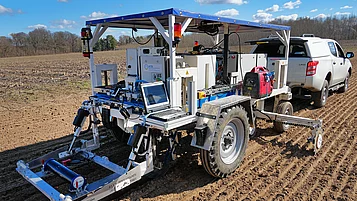 Ein landwirtschaftliches Fahrzeug steht mit einem Sensorsystem auf einem Anhänger auf einem Acker. Es hat große Reifen, eine Plattform mit einem Laptop und verschiedene technische Komponenten. Im Hintergrund ist ein Pickup zu sehen, der den Anhänger zieht. Der Himmel ist klar mit einigen Wolken.