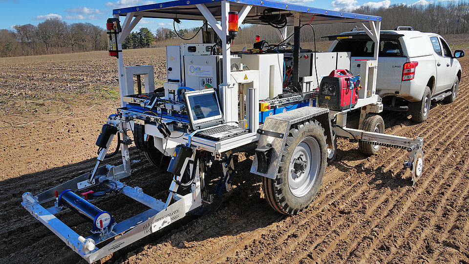 An agricultural vehicle with a sensor system is standing on a trailer in a field. It has large tires, a platform with a laptop and various technical components. A pickup truck can be seen in the background pulling the trailer. The sky is clear with some clouds. 