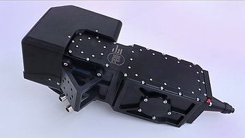 Black mechanical device with a rectangular top and a cylindrical outlet opening. It is equipped with screws and a labeled area.