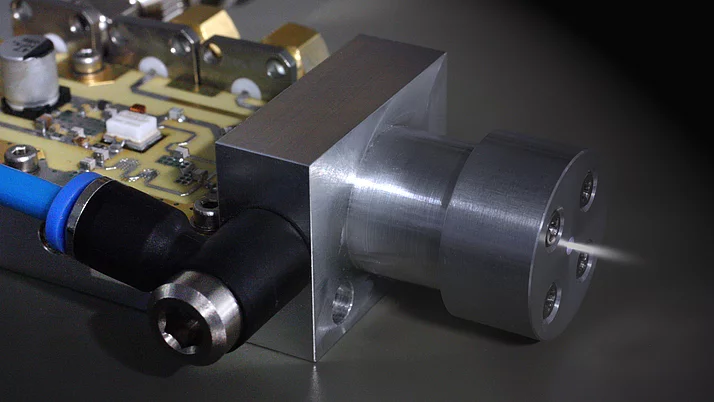 Close-up of a metallic component featuring a cylindrical nozzle emitting a fine mist. The base connects to a circuit board with various electronic elements.