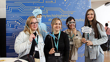 Vier Mädchen posieren mit einer Puppe in Reinraumkleidung vor einem blauen Banner mit technischen Motiven. Sie halten kreative Requisiten in der Hand. Die Atmosphäre ist freundlich und einladend.