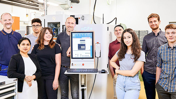 A diverse group of nine people stands together in a laboratory setting, smiling and posing for the camera. They are positioned in front of a computer workstation displaying software, with various laboratory equipment visible in the background.