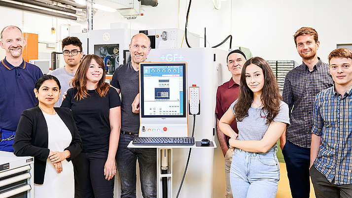 Eine Gruppe von acht Personen steht in einem Labor vor einem Computer. Die Umgebung ist hell und modern, mit verschiedenen Laborwerkzeugen im Hintergrund.