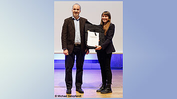 A man and a woman are standing on a stage. The man hands the woman a certificate. Both are smiling.