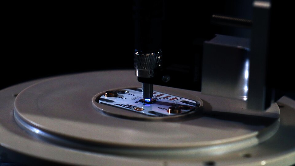 Eine Nahaufnahme eines präzisen Geräts, das eine elektronische Platine bearbeitet. Ein kleiner Mechanismus sitzt über der Platine und könnte entweder Löten oder Messen durchführen. Die Umgebung ist dunkel, wodurch der Fokus auf dem Gerät liegt.