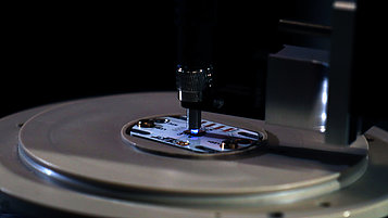 Close-up image of an LED in a lab mounted on a white subsmount. It emits a very small blue beam of light into a fiber above it. The holder rests on a round surface. The focus of the image is on the light and the contact between the LED and the fiber.
