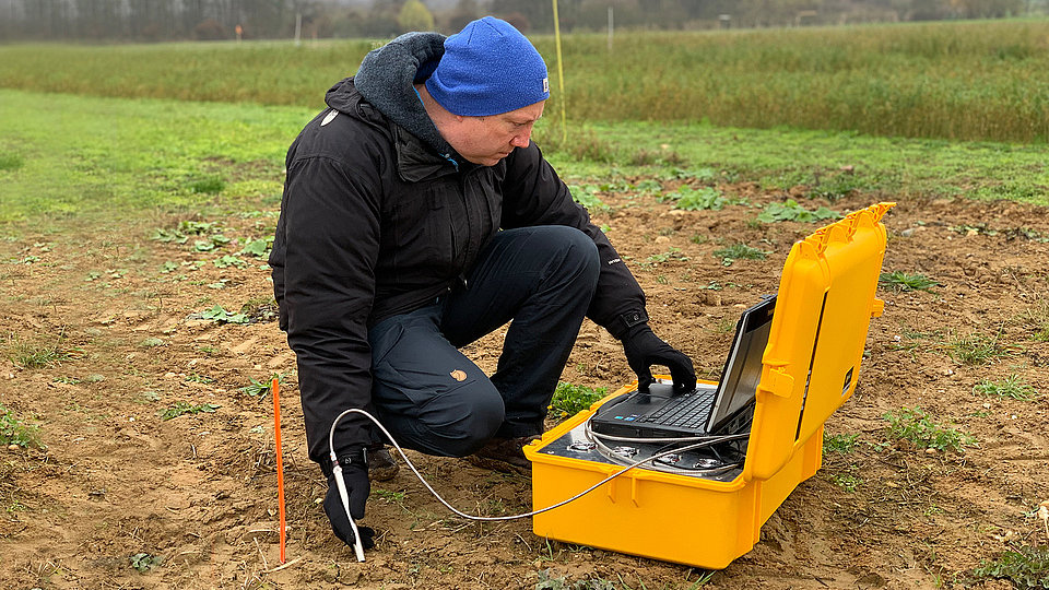 Man on knees in a field on the ground, measuring soil texture with a probe. The data is collected with a portable spectrometer in a case.