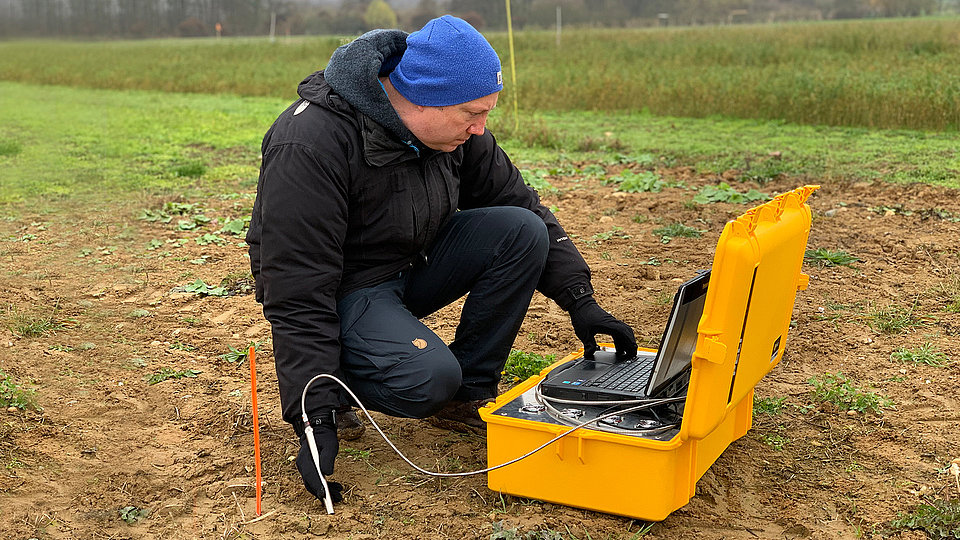 Mann kniet auf einem Feld und hält eine Messoptode in den Boden, um Raman-Messungen mit einem selbst entwickelten System durchzuführen