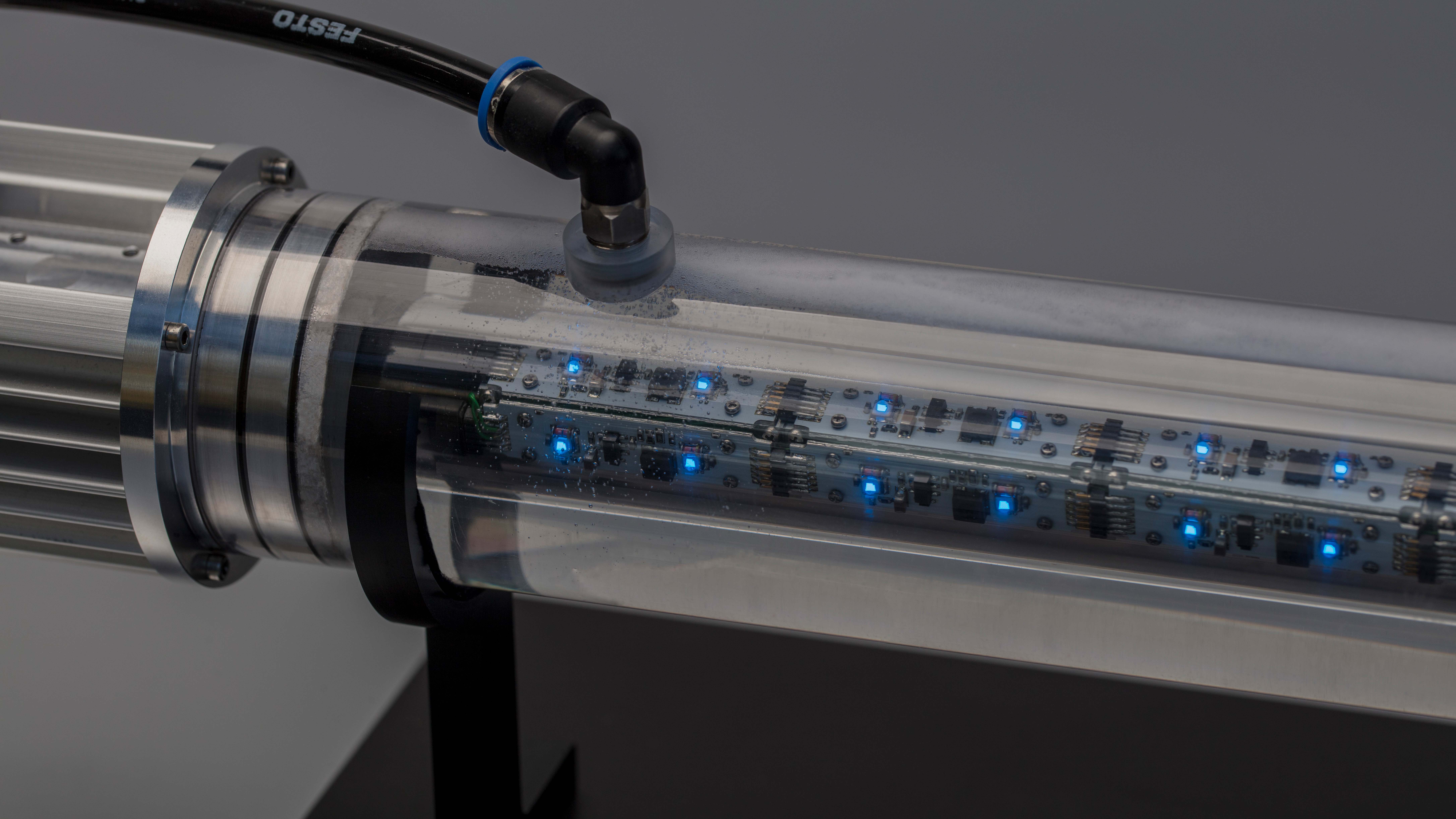 Transparenter Zylinder mit eingebauten, blauen LED-Leuchten und elektronischen Komponenten. An einem Ende des Zylinders ist ein Verbindungselement zu sehen. 